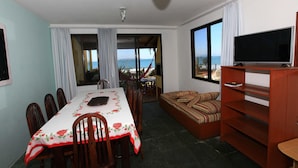 Une pièce comprenant une table à manger, des chaises, un canapé, un téléviseur et donnant sur la mer.