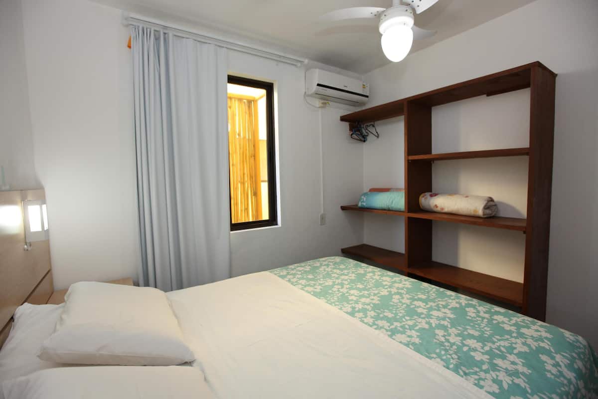 Un dormitorio con cama, ventana con cortinas, ventilador de techo y estante con toallas.