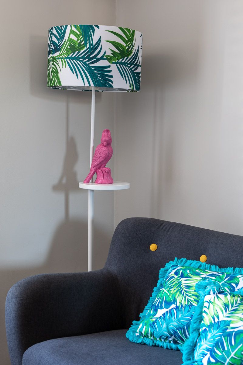 Een kamer met een grijze bank, een witte vloerlamp met een bladmotief, een roze vogelbeeldje op een witte plank, en een kussen met een blauw-groen bladpatroon.