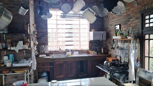 A rustic kitchen with a brick wall, wooden cabinets, and a window with lace curtains. - Sítio Sapiranga (Sapiranga)