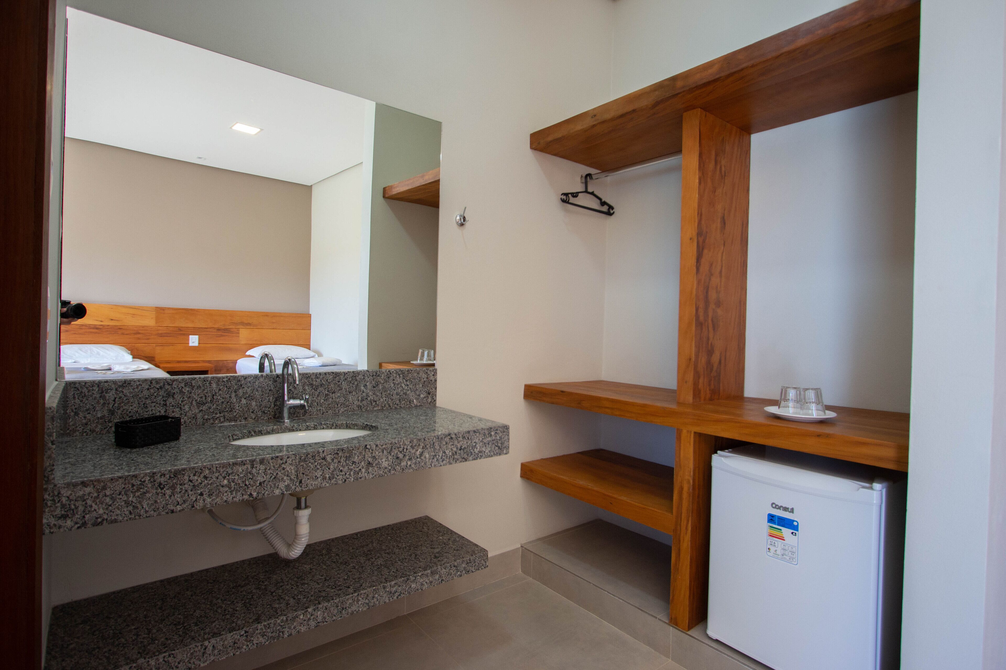 A bathroom with a granite countertop sink, a mirror, a wooden shelf, and a small refrigerator.