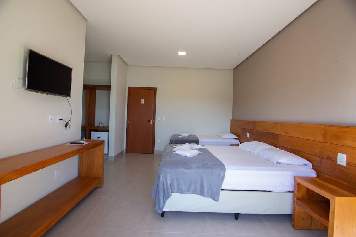 A hotel room with a large bed, a TV mounted on the wall, a wooden bench, and a door leading to another room.