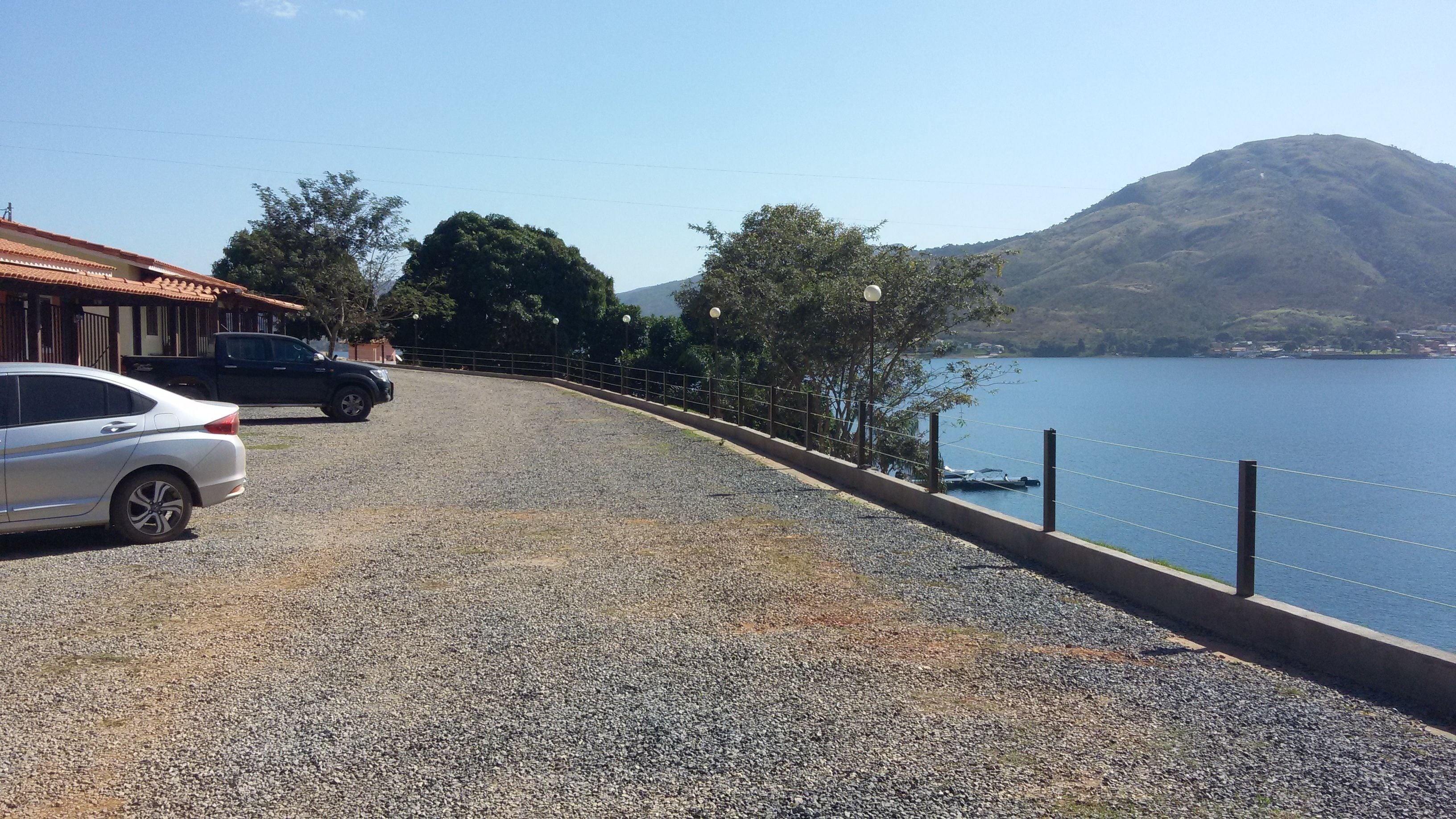 A gravel driveway leading to a building with a red-tiled roof, parked cars, and a view of a lake with a mountain in the background.