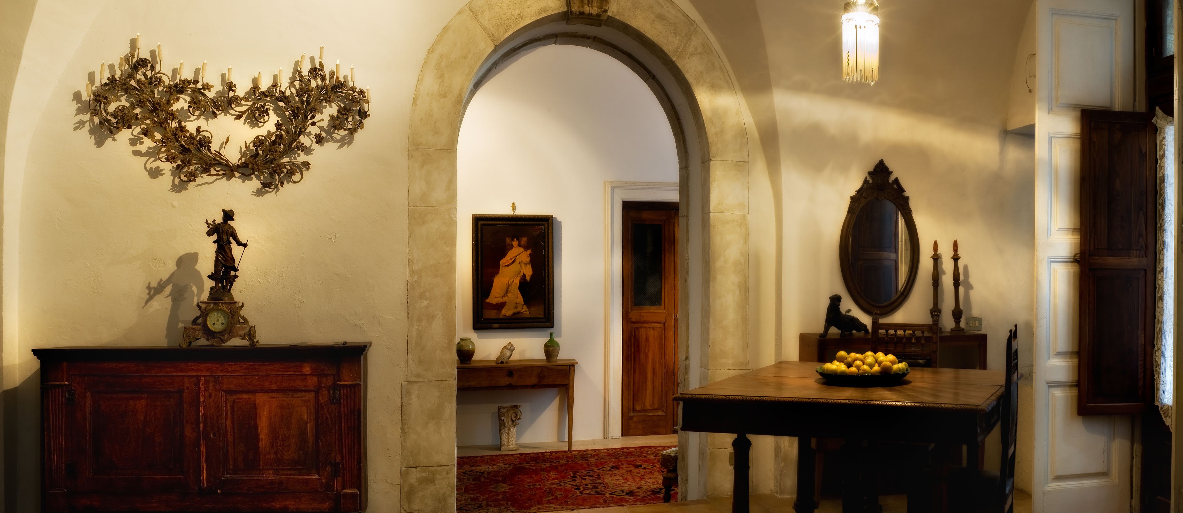 A room with a wooden cabinet, a wall-mounted clock, a chandelier, a framed painting, a wooden table with fruit, and a mirror.