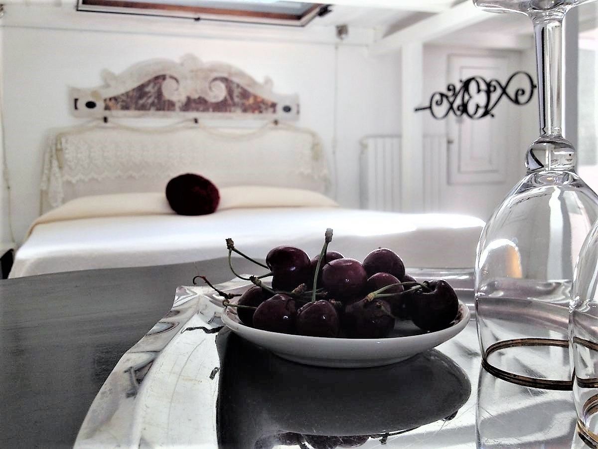 A bed with a white headboard and a decorative wall piece, a bowl of cherries on a table, and a clear glass bottle.