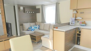 Et moderne, kompakt opholdsrum med sofa, et lille spisebord, et tekøkken med skabe og et vindue med gardiner.