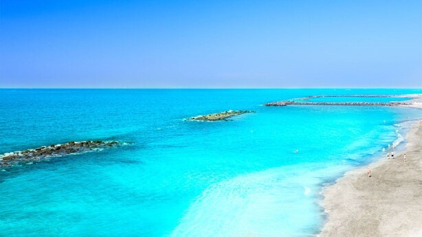Ein Strand mit klarem türkisblauem Wasser, einem Sandufer und einem steinernen Wellenbrecher.