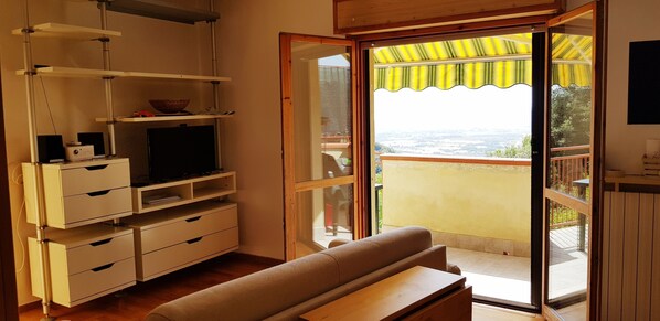 Uma sala de estar com televisão, armários e um sofá. Possui uma porta de vidro deslizante que dá para uma varanda com toldo amarelo.