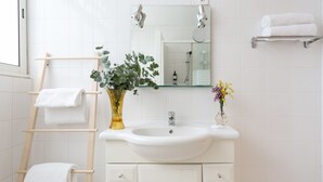 Et badeværelse med en hvid vask, et spejl over vasken, et håndklædetørrestativ med håndklæder, en vase med blomster og et bruseområde i baggrunden.
