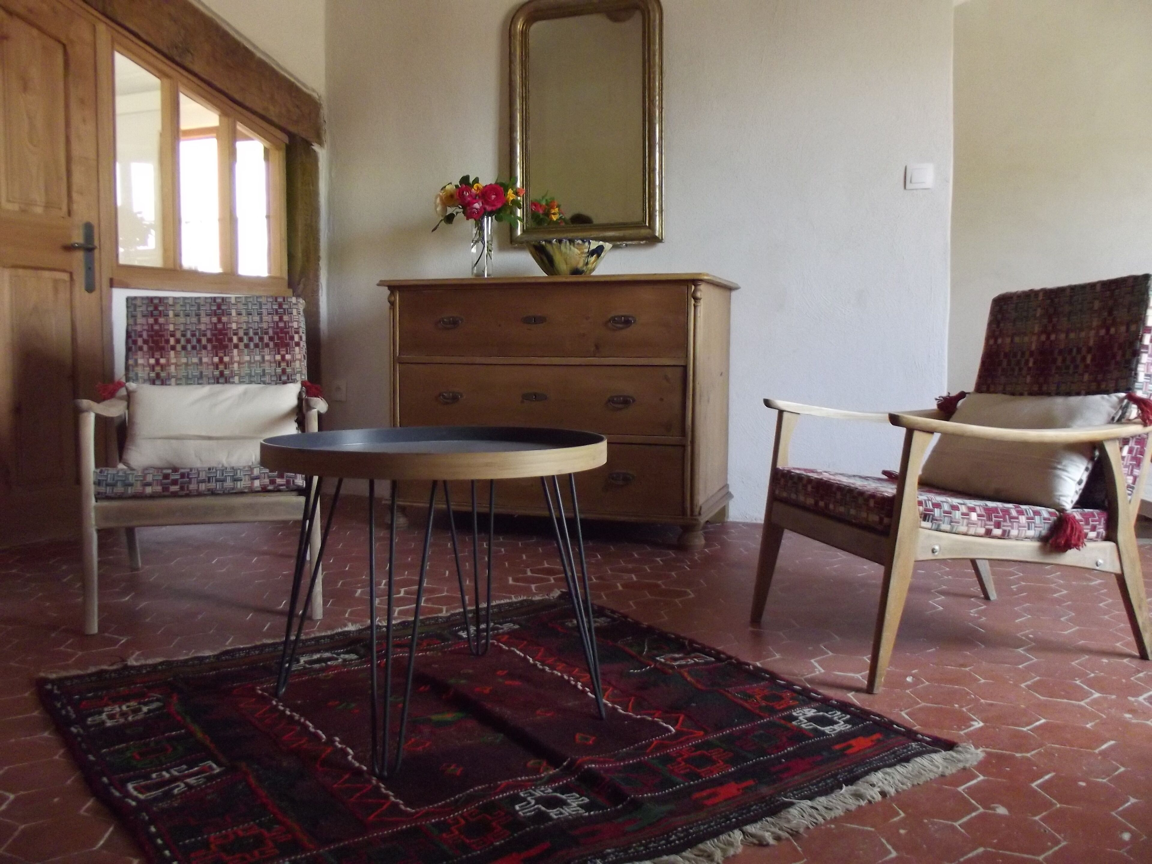 A room with a wooden dresser, two patterned chairs, a small round table, a mirror, and a red patterned rug on a brick floor.