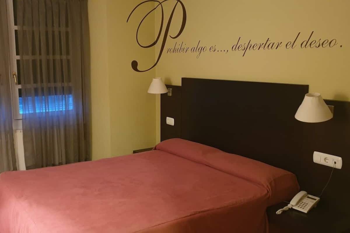 Quarto de hotel com cama, duas luminárias de cabeceira, telefone e parede com frase motivacional.
