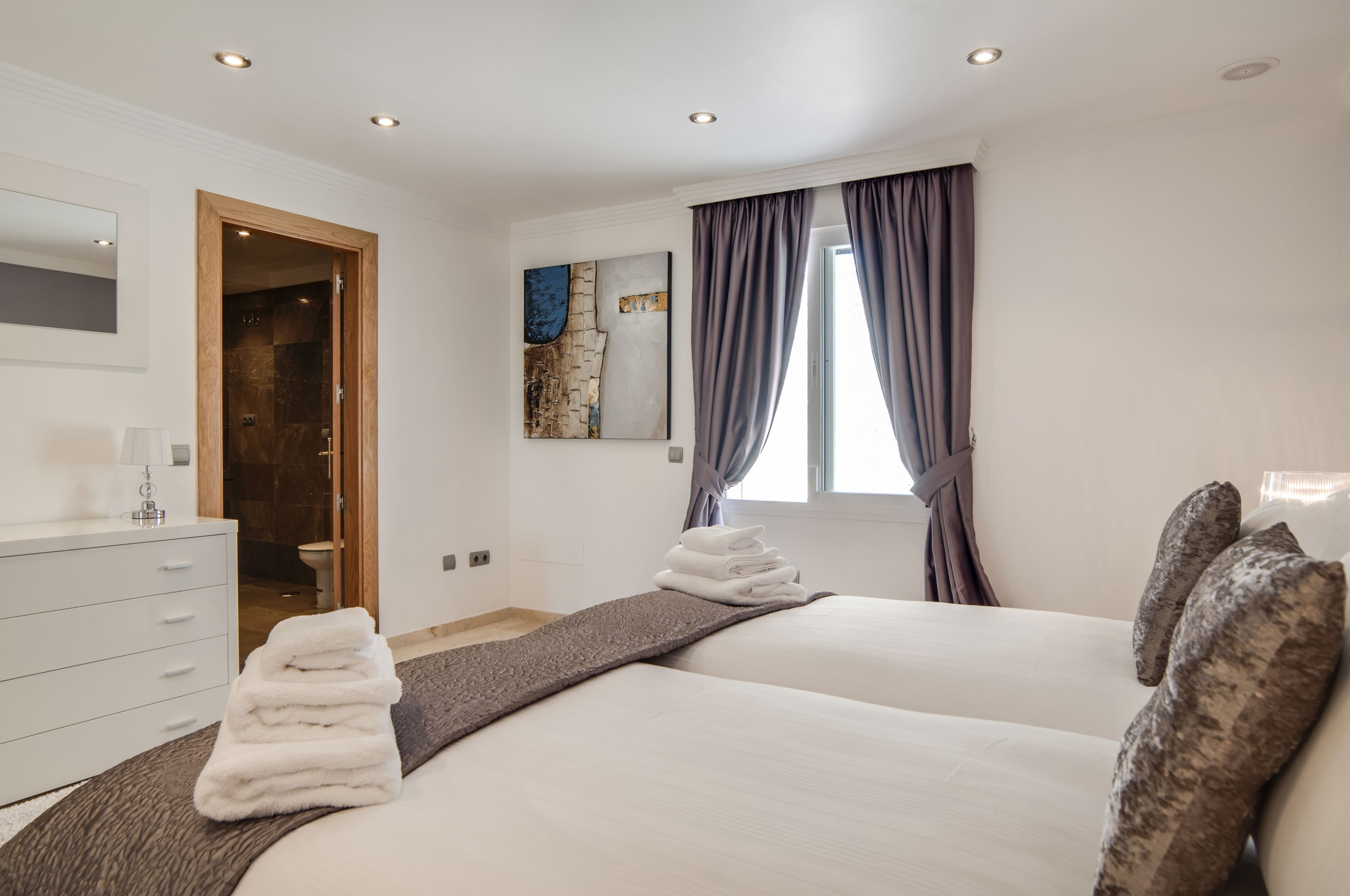 A modern hotel room with a large bed, a wooden door leading to a bathroom, a painting on the wall, and a window with curtains.