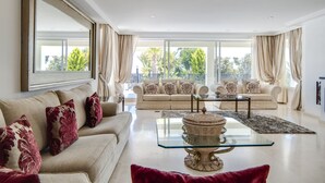 Amplia sala de estar con sofá color beige, una mesa de centro de vidrio y un ventanal con cortinas.