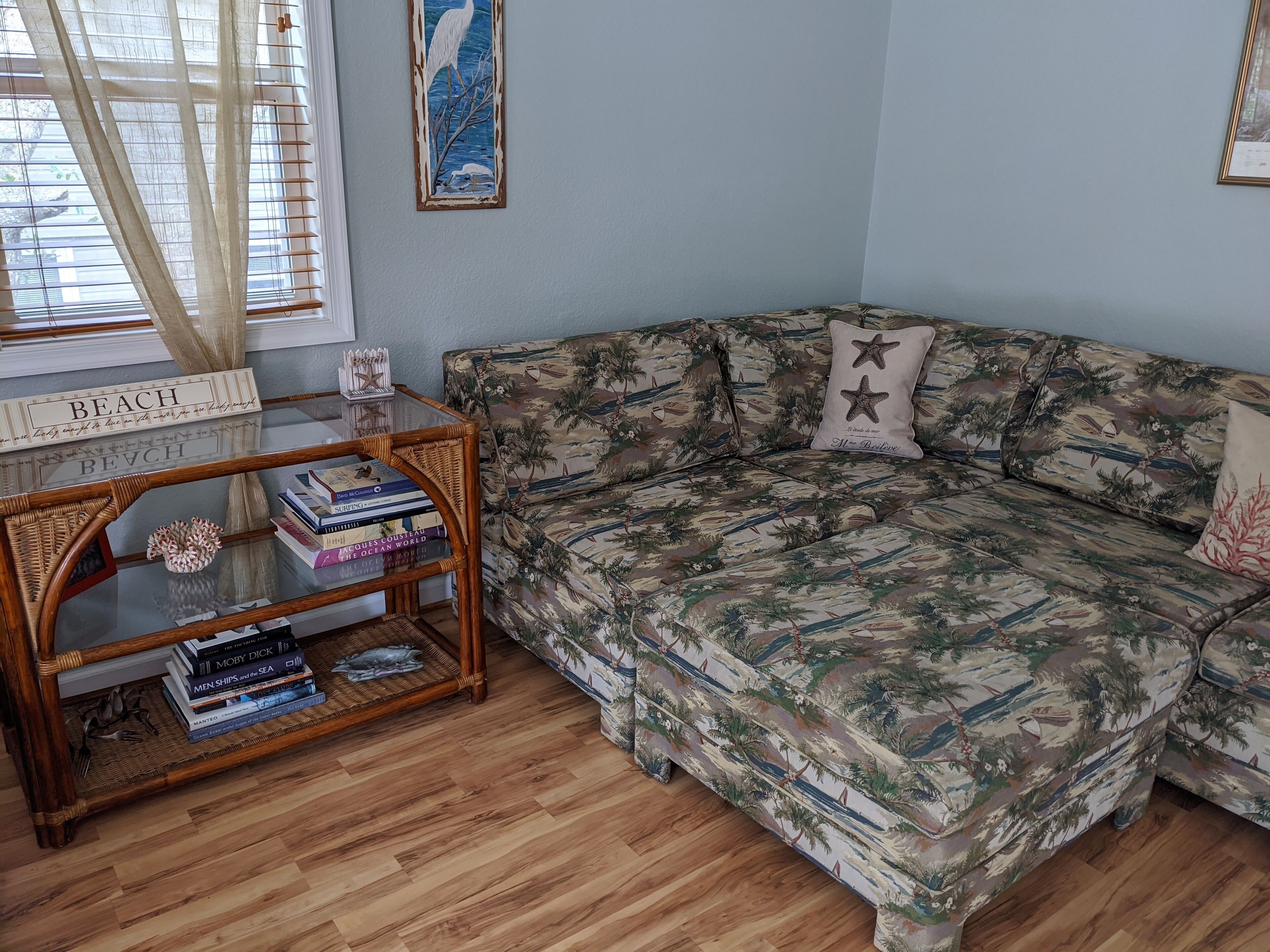 A living room with a patterned sofa, a glass coffee table with books, and a window with blinds.