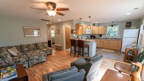 A spacious living room with a sofa, a coffee table, and a dining area with a table and chairs. The kitchen is visible in the background with wooden cabinets and a refrigerator.