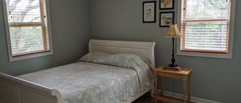 Un dormitorio con cama, dos ventanas con persianas, una mesita de noche con lámpara y una alfombra.