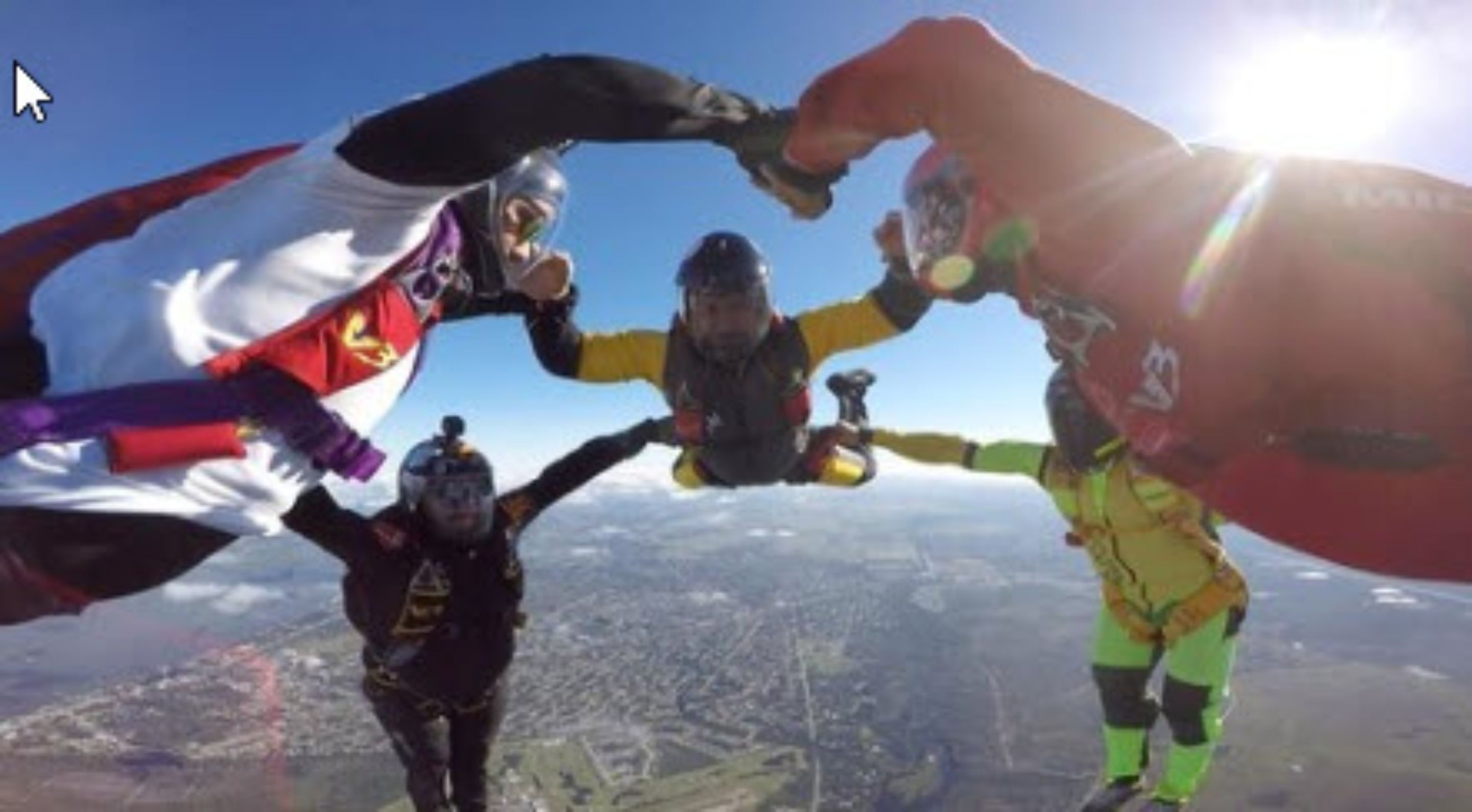 Three skydivers in mid-air with parachutes, one wearing a white and red flag, another in yellow and black, and the third in black and red.
