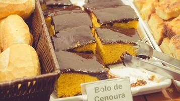 Un bufé con una variedad de productos horneados, incluyendo un pastel cubierto de chocolate identificado como "Bolo de Cenoura".