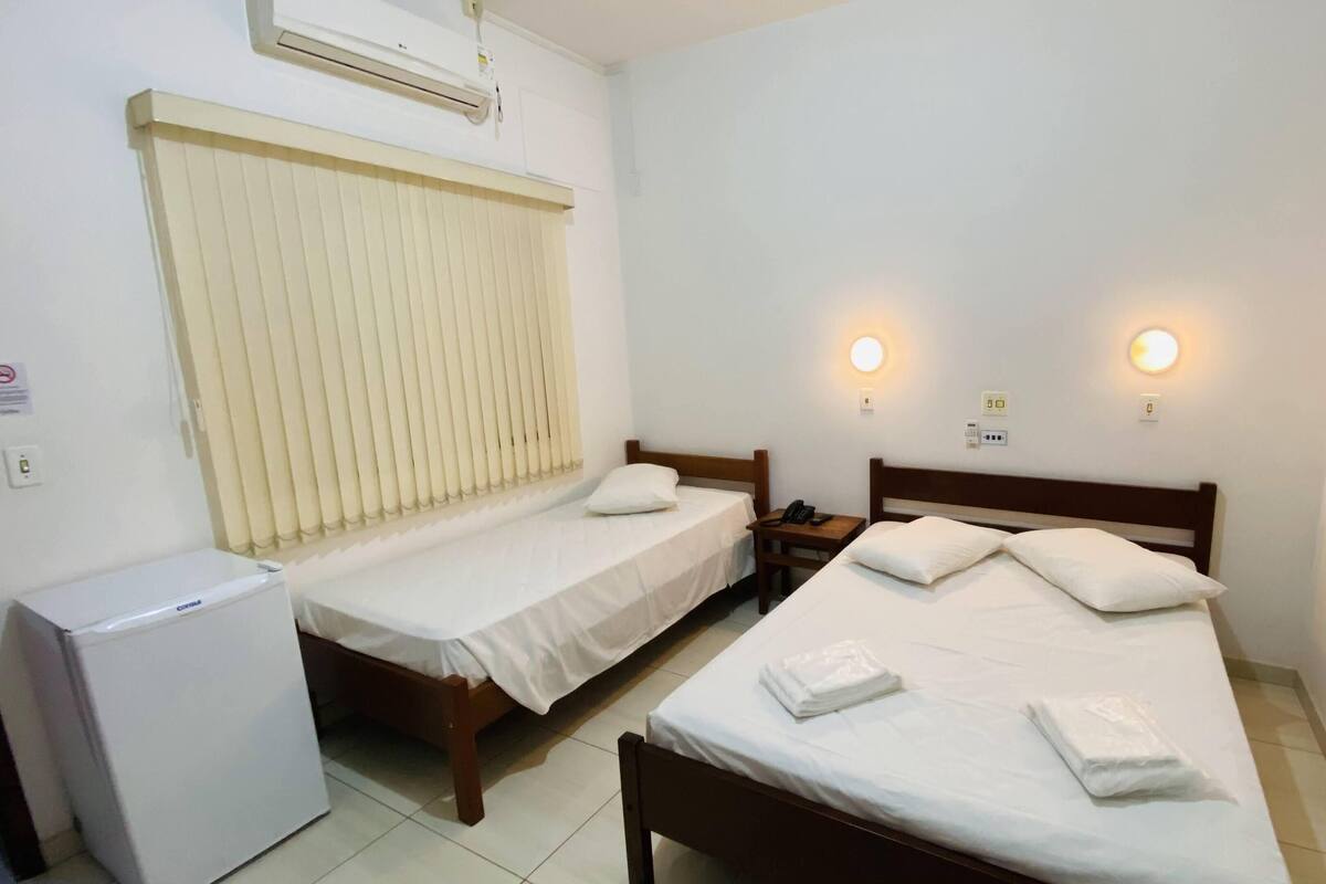Ein Hotelzimmer mit zwei Einzelbetten, einem kleinen Nachttisch mit Telefon, einer wandmontierten Klimaanlage und einem Fenster mit Jalousien.