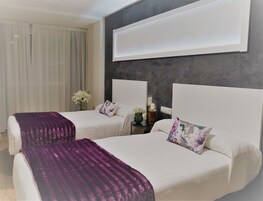 Habitación de hotel con dos camas, un televisor grande montado en la pared y una mesita de noche con un florero y una vela.