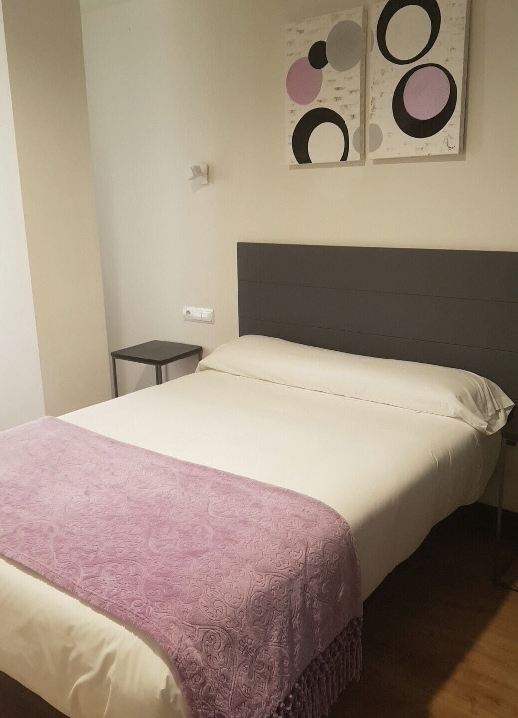 Una cama bien tendida con un cabecero gris, una mesita de noche pequeña y una pared con arte abstracto.
