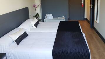A hotel room with two beds, a bedside table, a small table with towels, a potted plant, and a door.