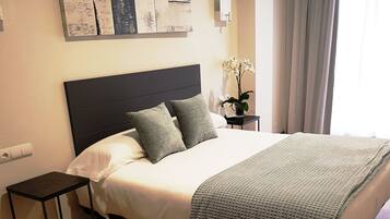 Habitación de hotel ordenada con una cama grande, una mesita de noche, una lámpara de pared y una ventana con cortinas.
