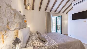 A bedroom with a stone wall, wooden beams, a bed with patterned bedding, a hanging light fixture, and a TV on the wall.