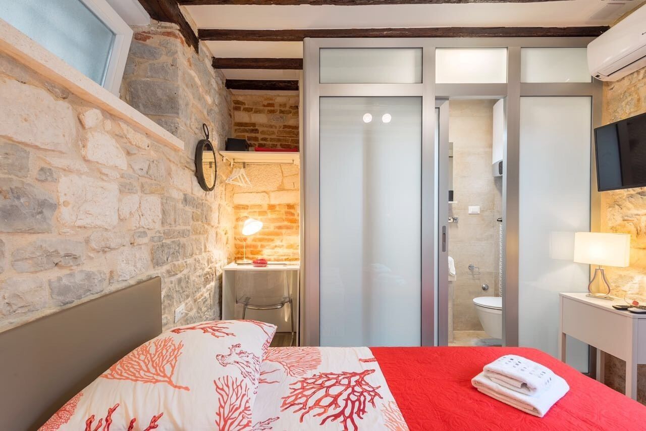A bedroom with a stone wall, a bed with a red blanket, a desk with a lamp, and a sliding door leading to a bathroom.