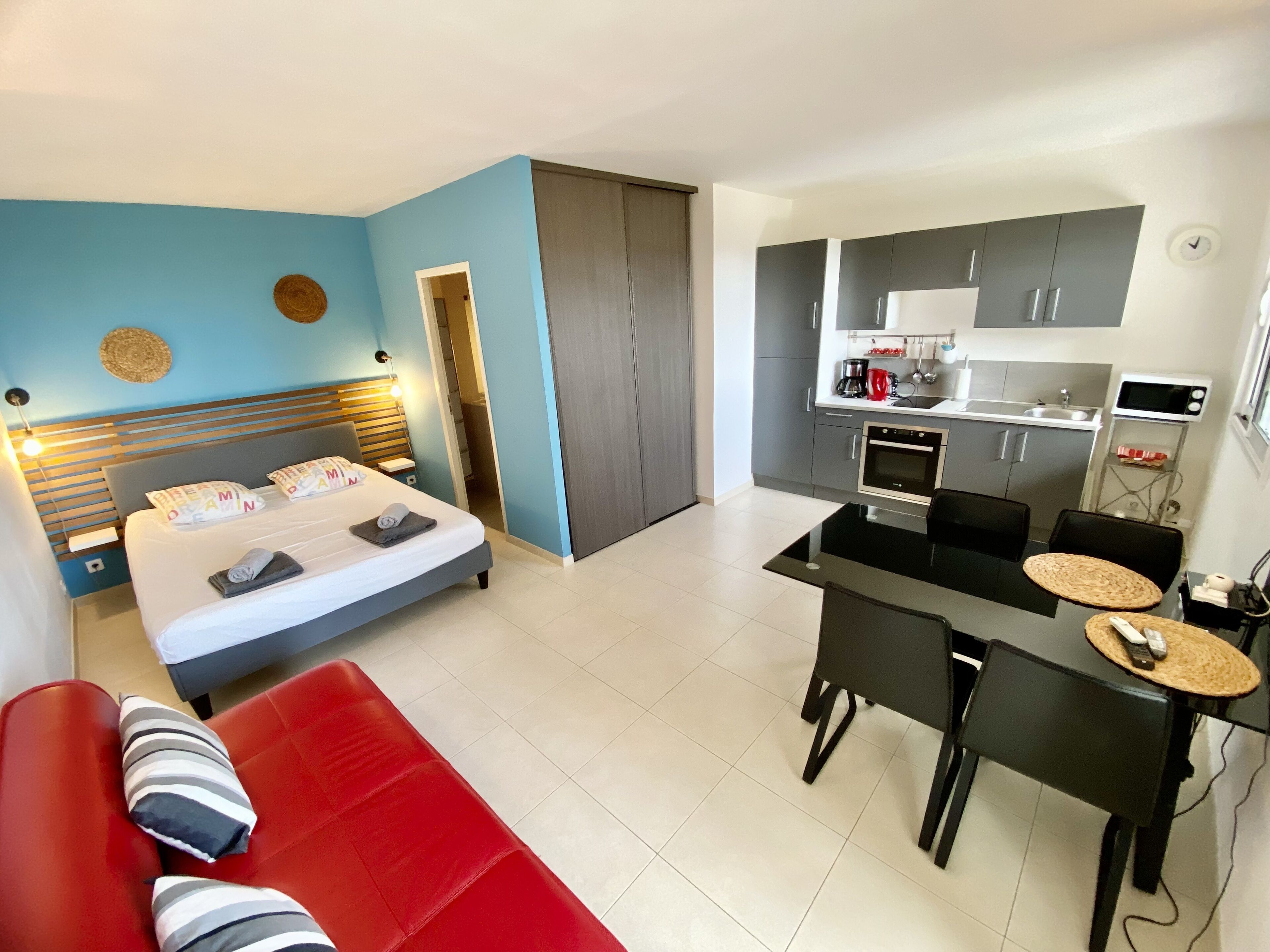 A modern hotel room with a bed, a red sofa, a dining area with a table and chairs, and a kitchenette.
