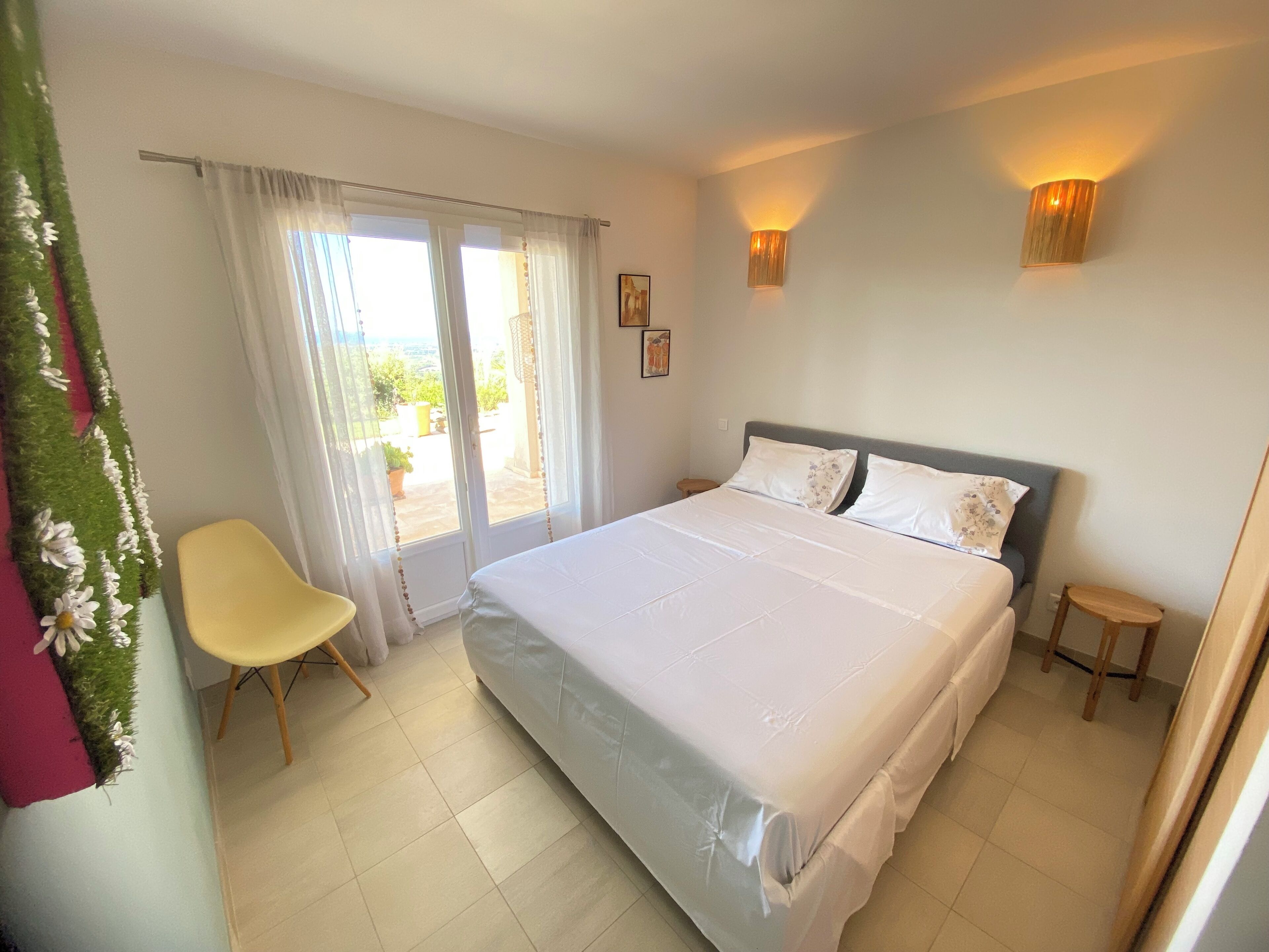 Une chambre à coucher avec un grand lit, une chaise, une petite table et une vue sur l’extérieur par une fenêtre avec des rideaux.