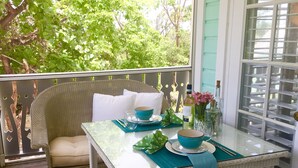 Une chaise en osier avec des coussins blancs, placée sur un balcon où se trouve une table en verre agrémentée d’un saladier bleu, d’une bouteille de vin et d’un vase de fleurs.