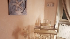 Ein Zimmer mit einem weißen Tisch und Stuhl, einem Kunstwerk mit Seestern an der Wand, einem Heizkörper und einem Fenster mit Vorhängen.