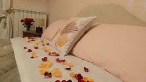 Ein ordentlich bezogenes Bett mit Rosenblättern auf der Bettdecke, eine Vase mit roten Rosen auf einem Nachttisch und eine Lampe.