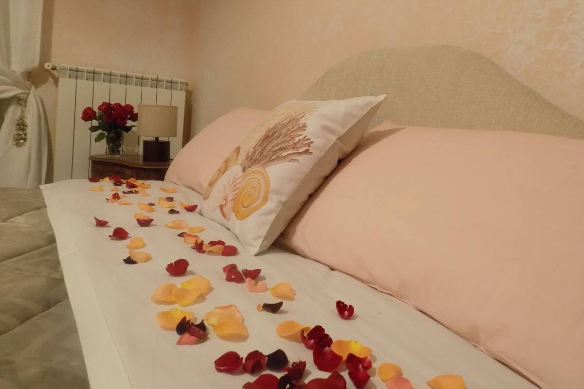A neatly made bed with rose petals scattered across the bedspread, a vase with red roses on a bedside table, and a lamp.