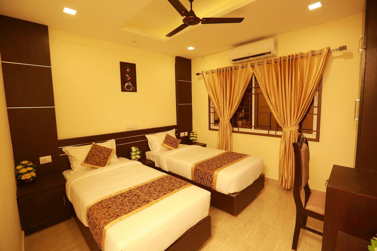 A hotel room with two beds, a ceiling fan, an air conditioning unit, and a window with curtains.