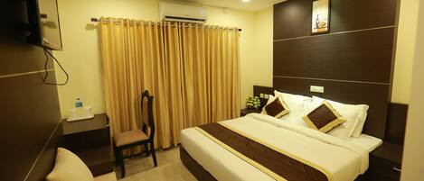 A hotel room with a large bed, a ceiling fan, an air conditioning unit, a chair, a small table with a water bottle, and a nightstand with a lamp.