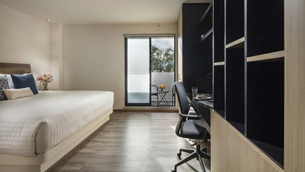 A modern bedroom with a large bed, a desk with a chair, and a sliding glass door leading to a balcony. - ROOM Hotel & Suites (Puebla)