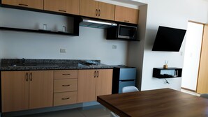 A modern kitchen with wooden cabinets, a granite countertop, a microwave, and a flat-screen TV mounted on the wall. - ROOM Hotel & Suites (Puebla)