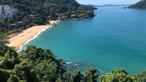 Une zone côtière avec une plage de sable, une mer d’un bleu limpide et une colline boisée.