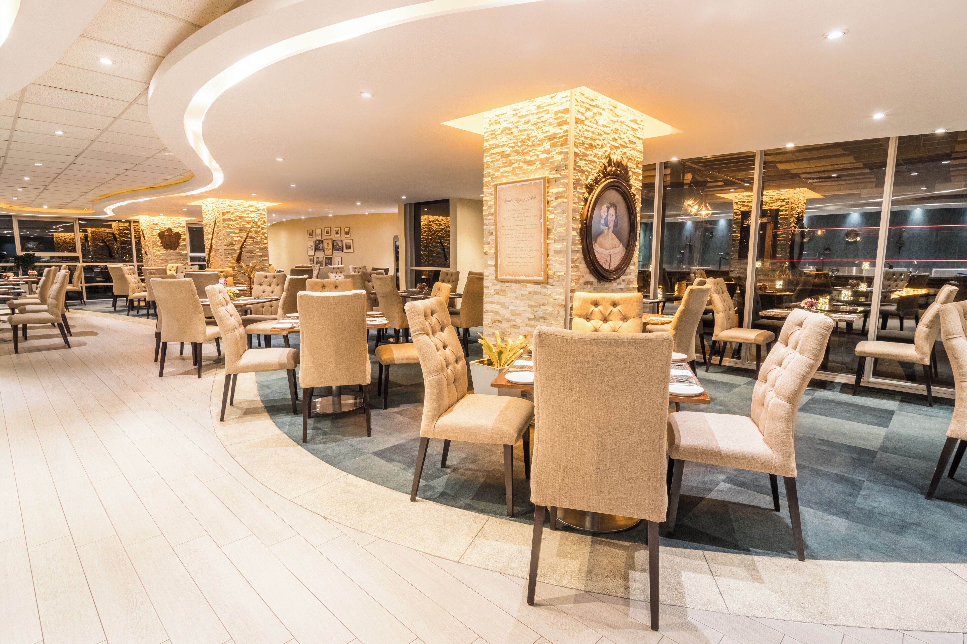 A modern restaurant with round tables and cushioned chairs, stone accents, and large windows.