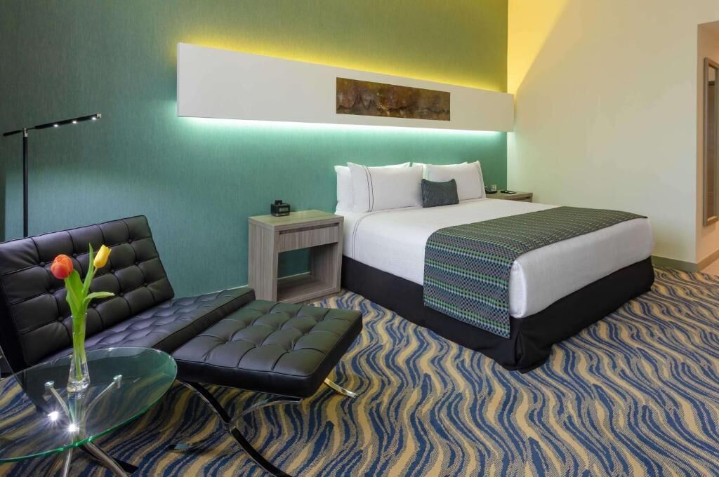 Une chambre d’hôtel avec un lit, une petite table sur laquelle se trouve un vase, une chaise et une lampe sur pied.