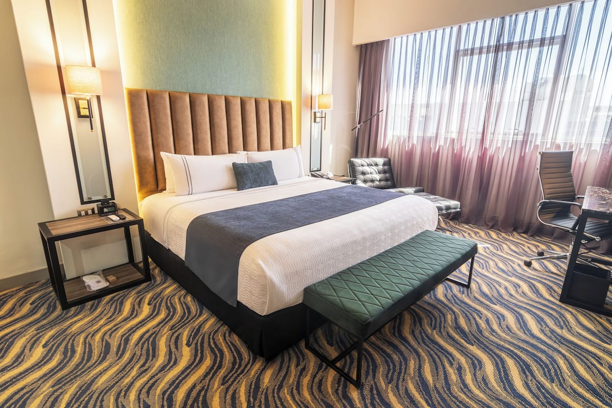 A hotel room with a large bed, a bench, a chair, a nightstand, and a view of the outdoors through curtains.