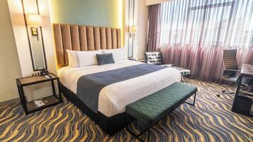A hotel room with a large bed, a bench, a chair, a nightstand, and a view of the outdoors through curtains.