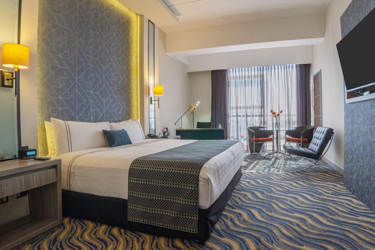 A hotel room with a bed, bedside table, lamp, desk, chair, and a TV mounted on the wall.
