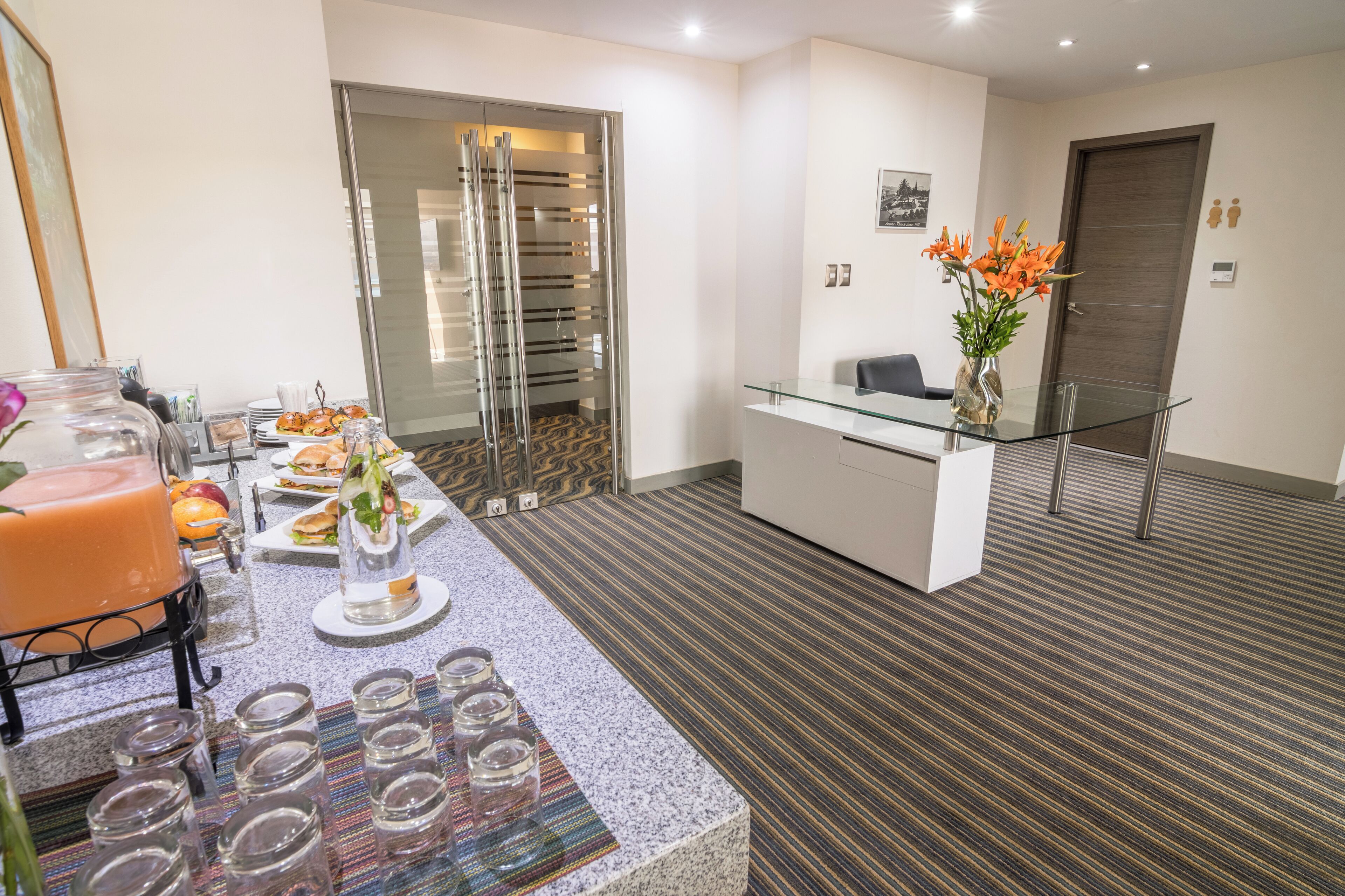 Un vestíbulo de hotel moderno con un mostrador de recepción, un separador de vidrio y una zona de bufé con comida y bebidas.