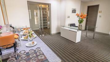 Un vestíbulo de hotel moderno con un mostrador de recepción, un separador de vidrio y una zona de bufé con comida y bebidas.