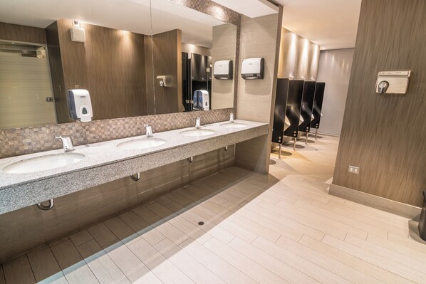 Un baño moderno con tres lavamanos, un espejo grande, secamanos y suelo de baldosas.