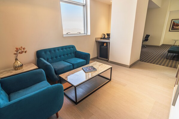 Una sala de estar moderna con un sofá azul, una mesa de centro de vidrio y una mesita auxiliar con un jarrón. Se ve una ventana que deja entrar luz natural y una zona de kitchenette con una cafetera.