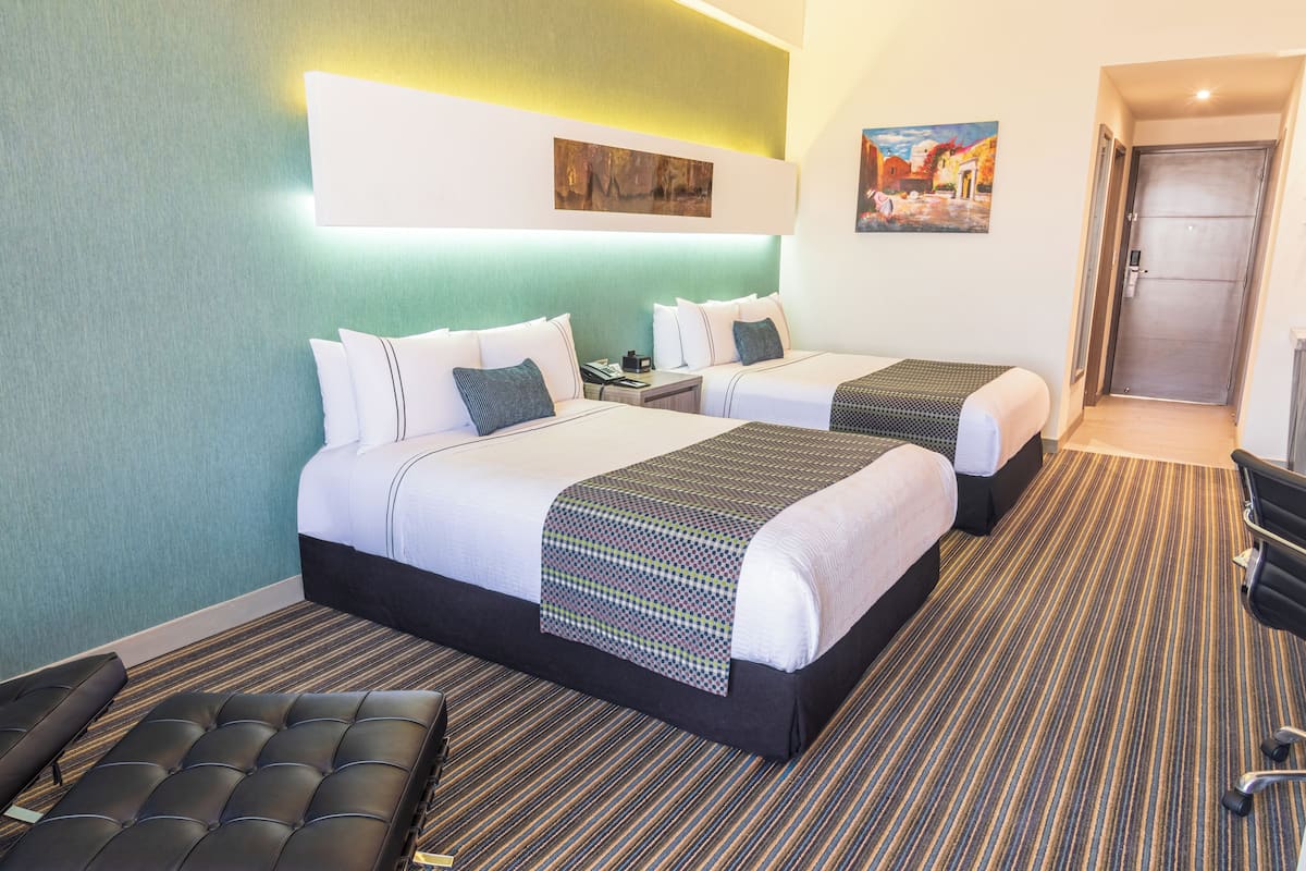 A hotel room with two beds, a bench, a desk, and a door.